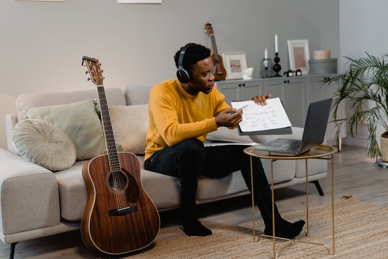 A man in a yellow sweater teaches guitar online using a laptop from his comfortable living room.